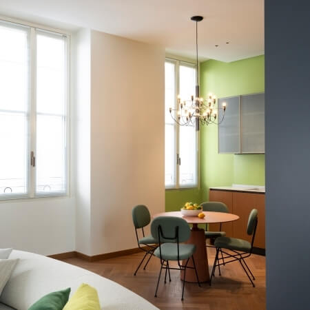 Modern dining room with a round table, green chairs, chandelier, and natural light from a large window.