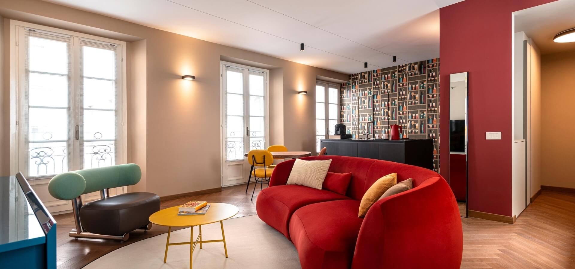 Modern living room with a red sofa, yellow coffee table, blue desk, and a cozy kitchen area, filled with natural light.