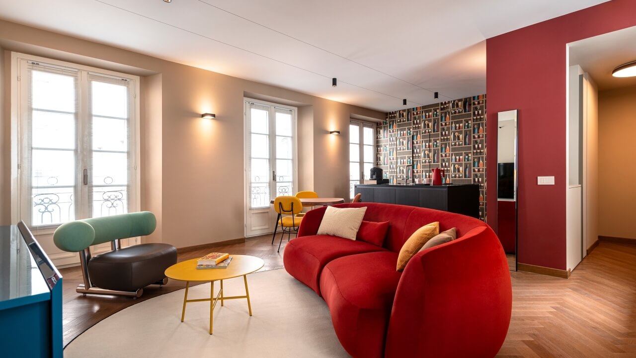 Modern living room with a red couch, yellow coffee table, green chair, wooden floor, and large windows for natural light.