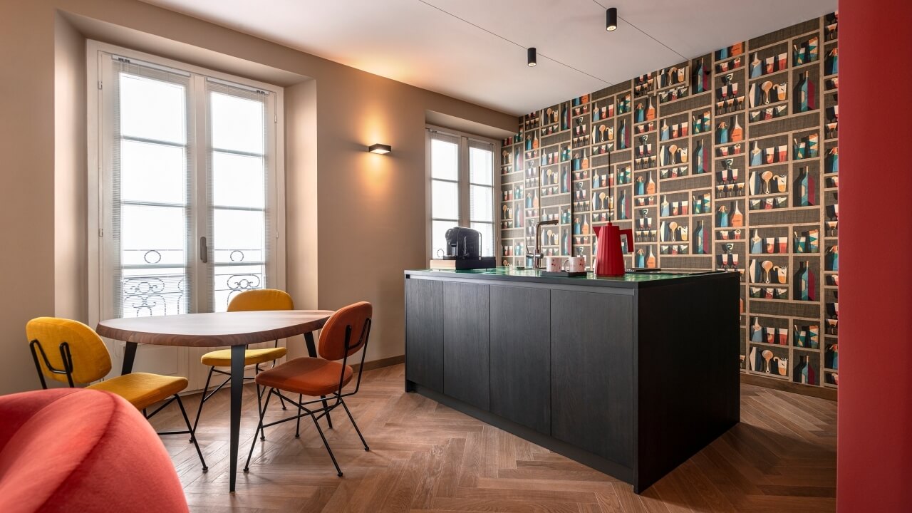 Modern kitchen with a round table, colorful chairs, central island, and a bookshelf wall, illuminated by natural light.