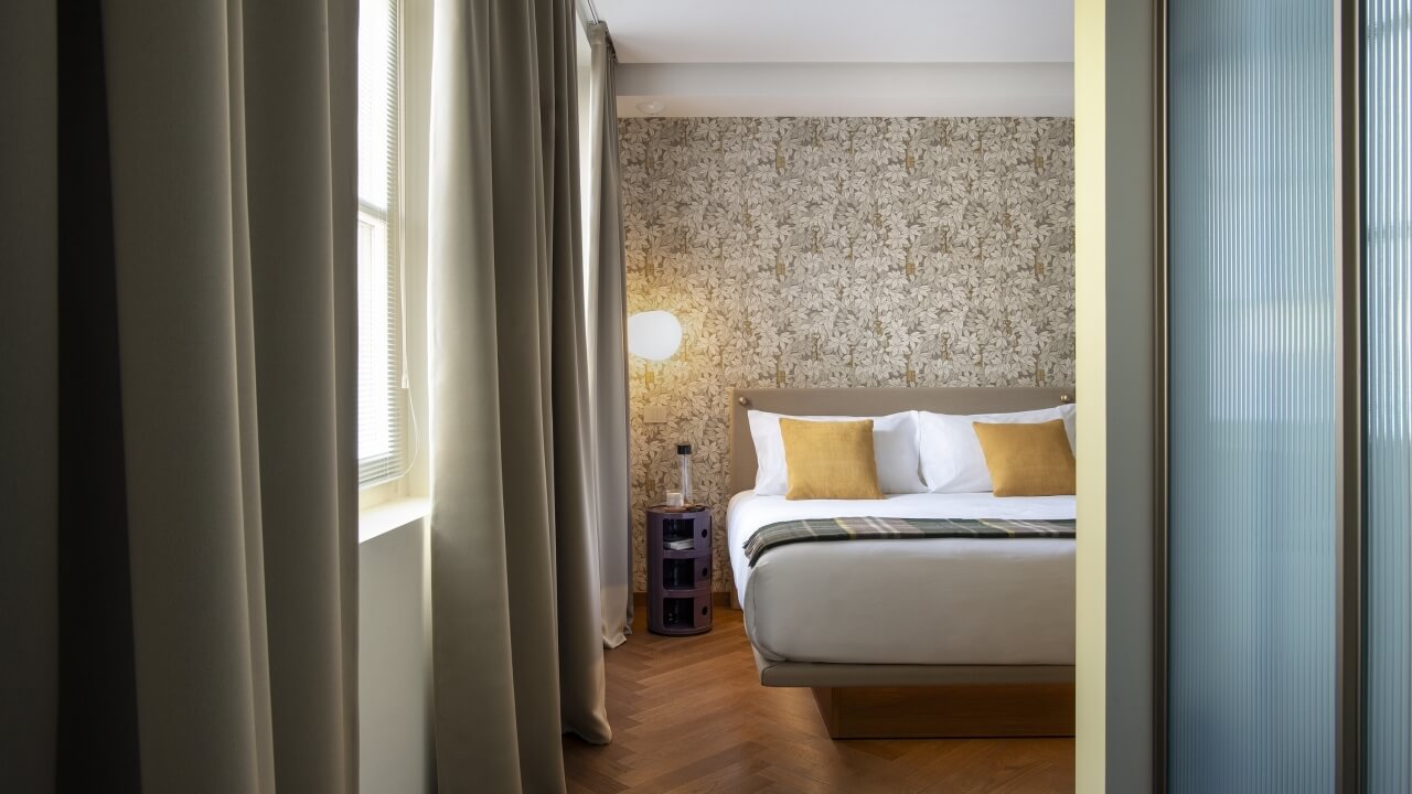 Serene bedroom with a floral wallpaper, white comforter, yellow pillows, nightstand, and warm wooden floor.