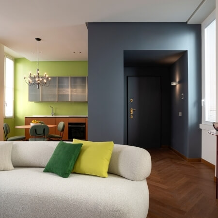 Modern living room with a white couch, vibrant pillows, eclectic furniture, sage green walls, and a compact kitchen area.