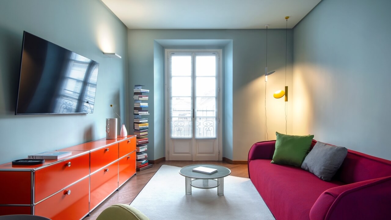 Modern living room with blue walls, a red couch, orange cabinet, and serene decor, featuring natural light and pastel colors.