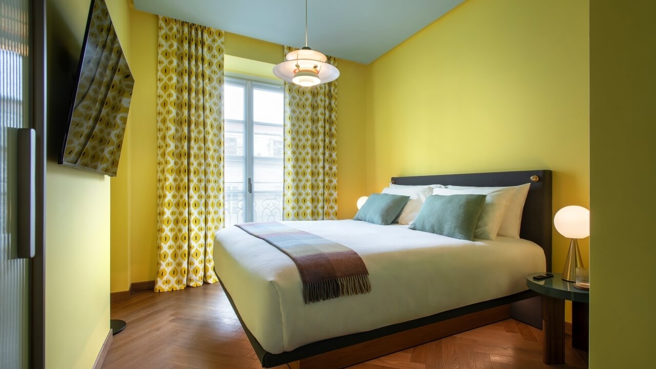 A bright bedroom with a bed, nightstand, lamp, and window with yellow curtains, featuring light wood flooring and blue ceiling.