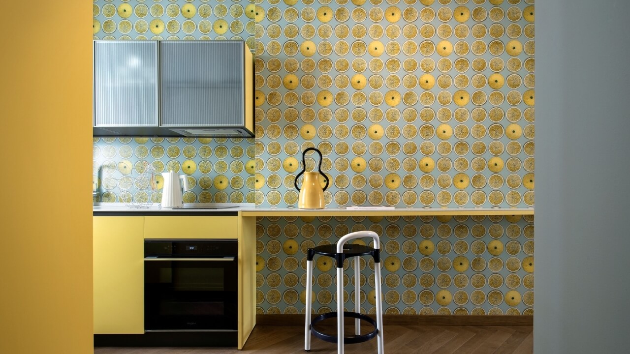 Bright kitchen with yellow cabinets, circular patterned wallpaper, oven, sink, and stool, creating a cheerful atmosphere.