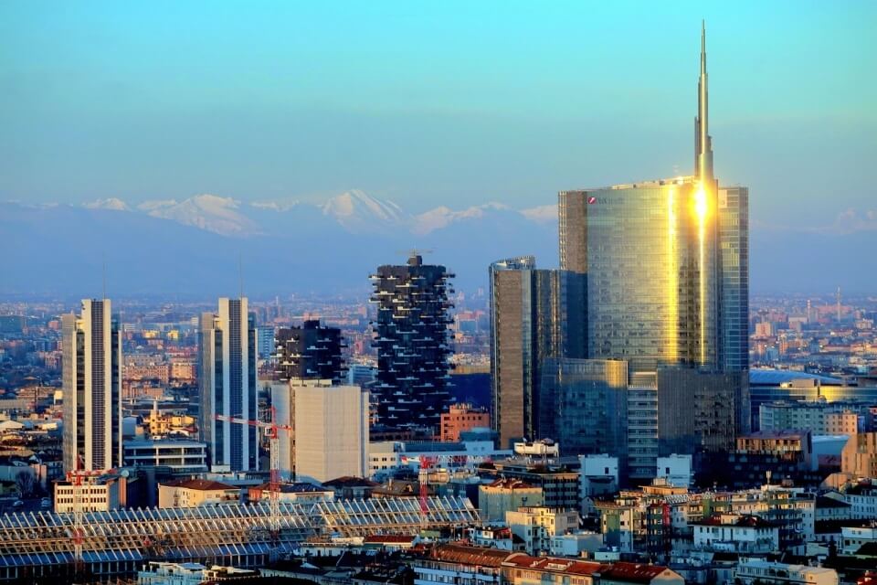 Cityscape featuring a prominent glass skyscraper, diverse buildings, and snow-capped mountains in the background.