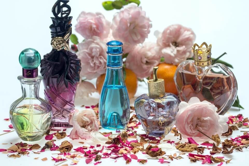 Disposizione di bottiglie di profumo con fiori rosa e petali, su sfondo bianco, con arance e rose sfocate sullo sfondo.