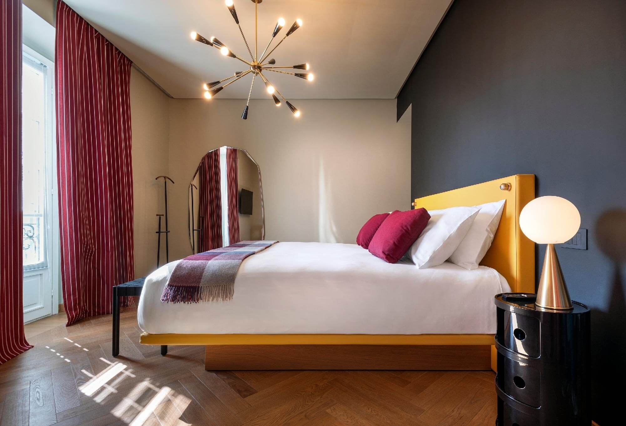 Stylish bedroom with yellow bed, starburst light fixture, red curtains, and dark wood flooring, exuding modern sophistication.