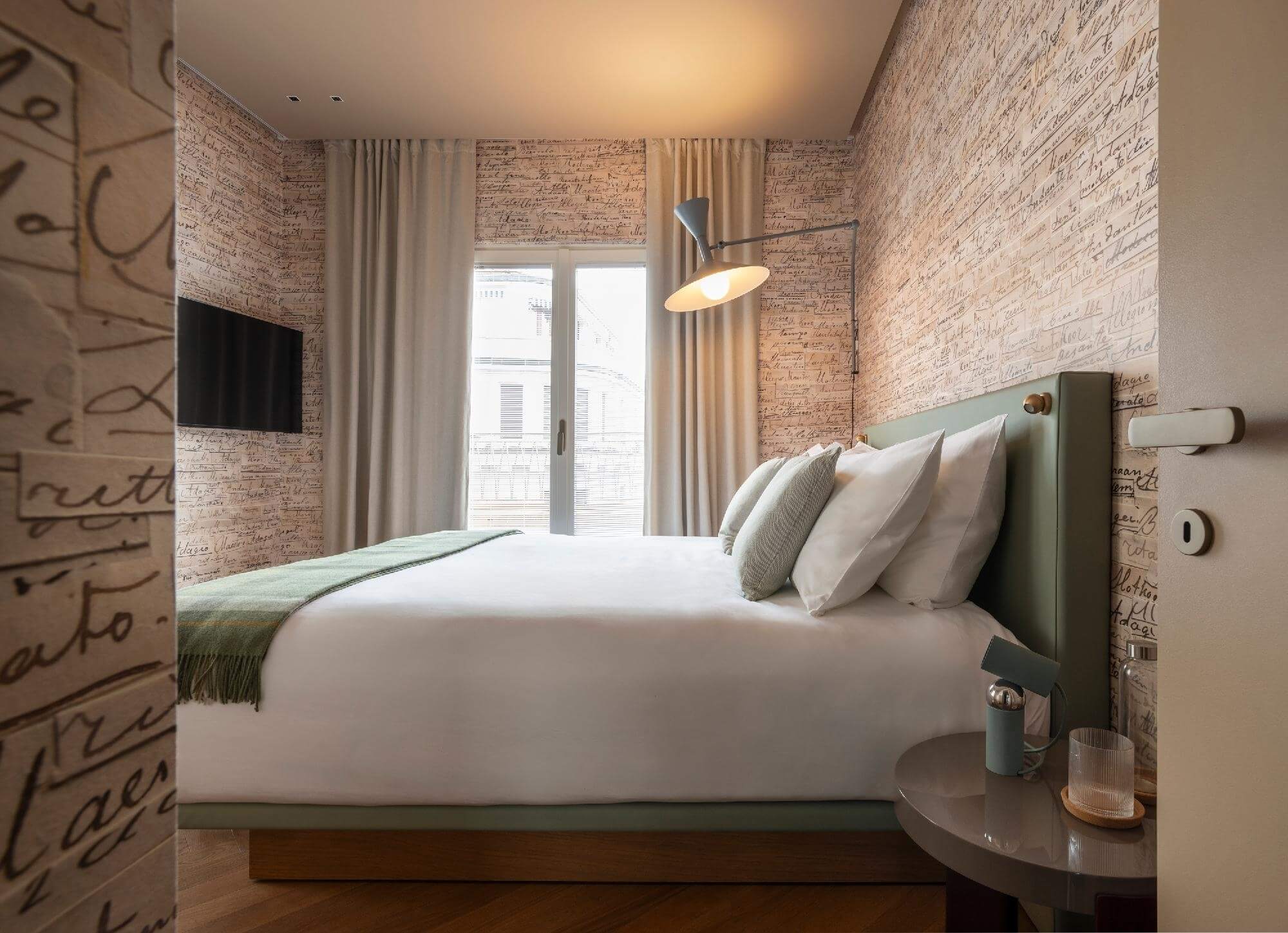 Camera da letto moderna e minimalista con letto verde, comforter bianco, tavolo grigio e vista su un edificio urbano.