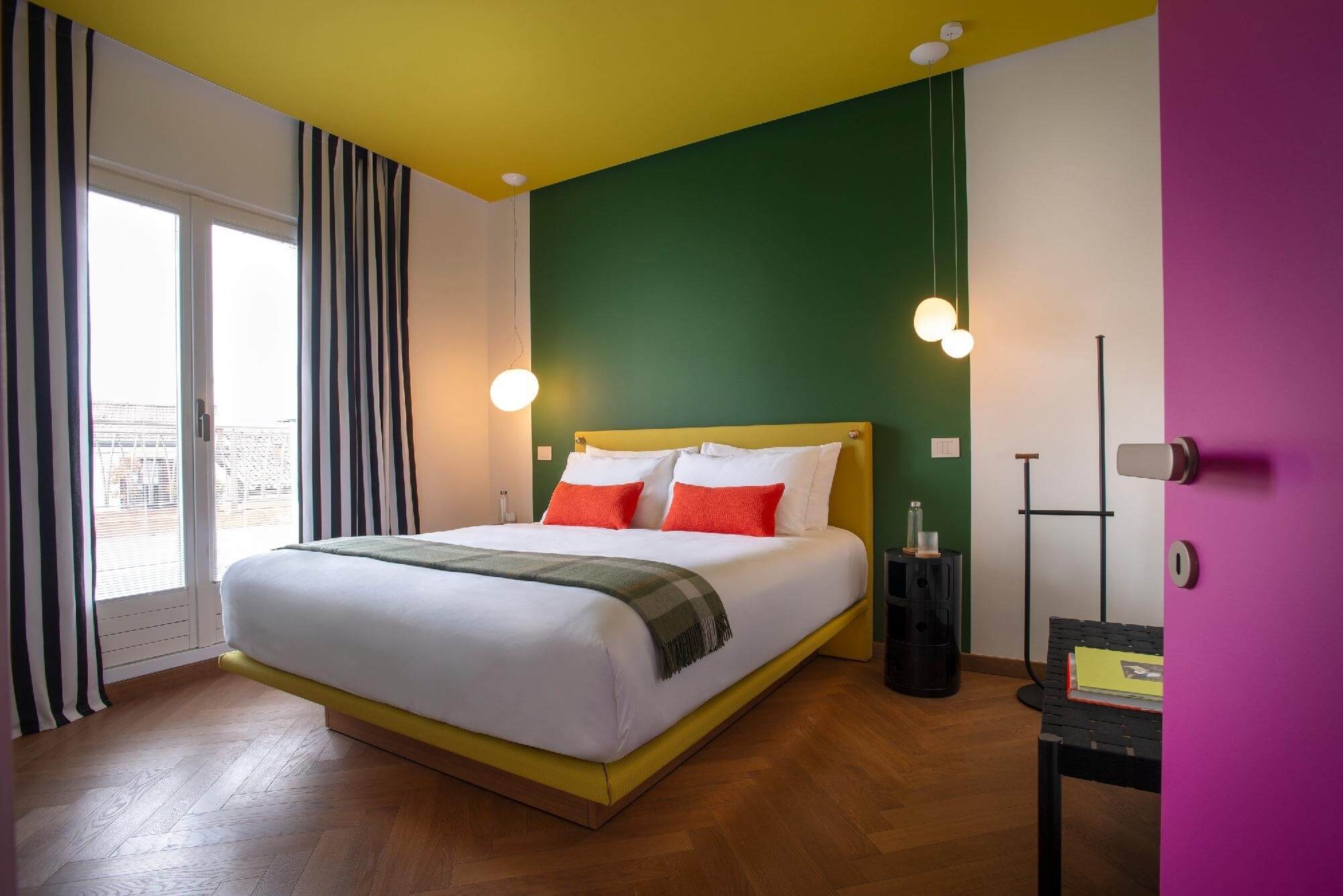 Cozy bedroom with a yellow bed, colorful pillows, hanging lights, striped curtains, and wooden herringbone flooring.