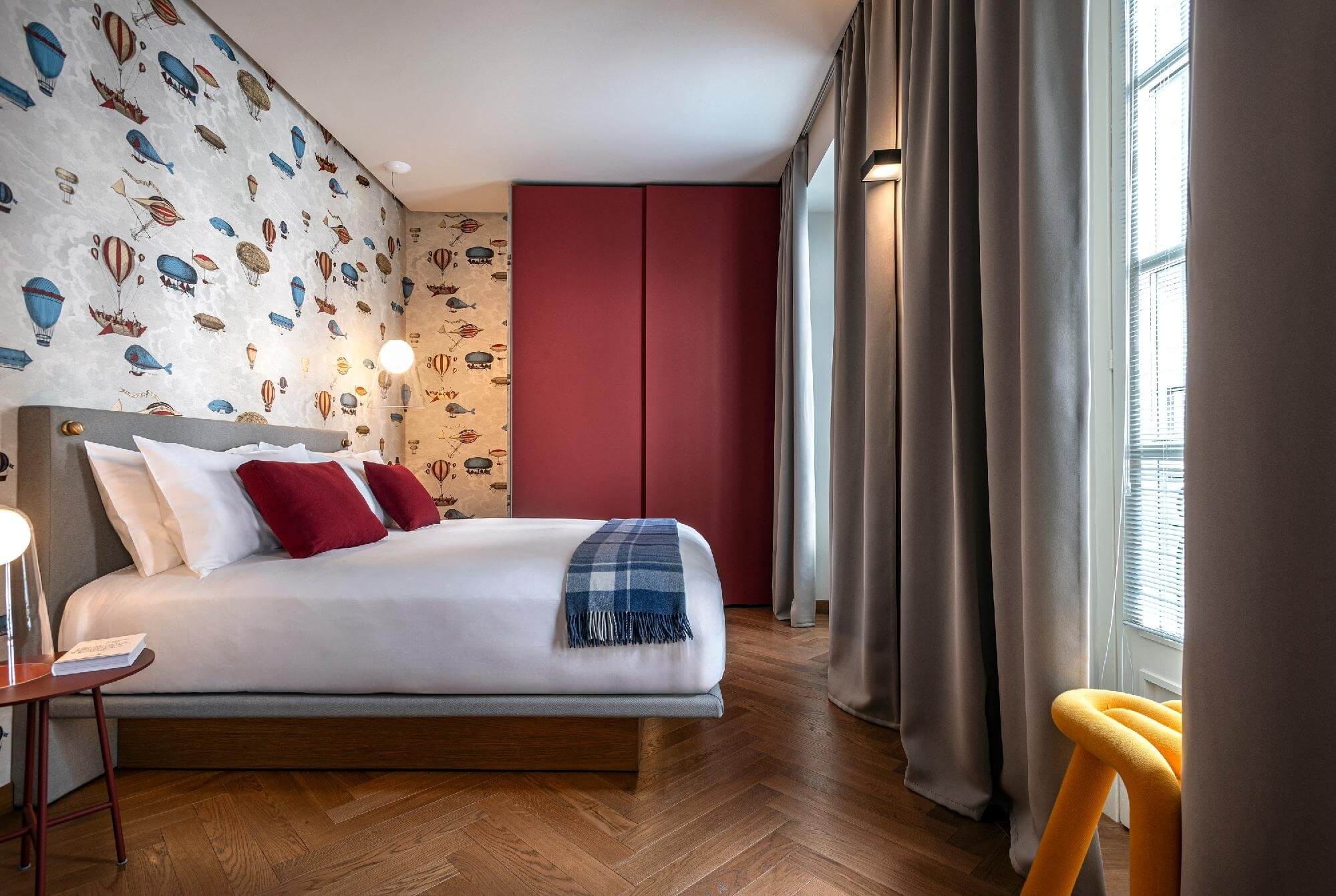 Modern bedroom with white theme, wooden accents, gray headboard, red pillows, whimsical wall patterns, and yellow chair.