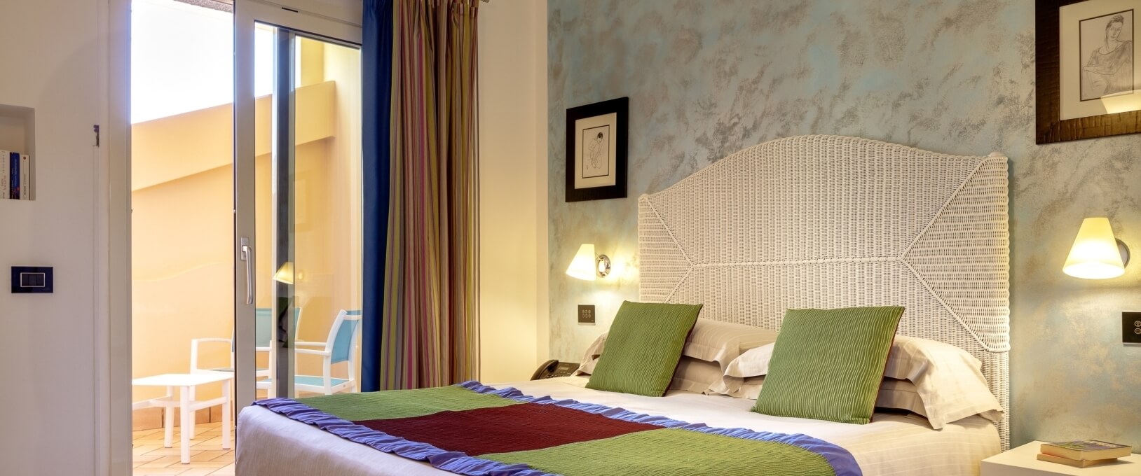 Chambre avec lit, table blanche, tableaux au mur, et coin salon extérieur visible à travers une porte vitrée.