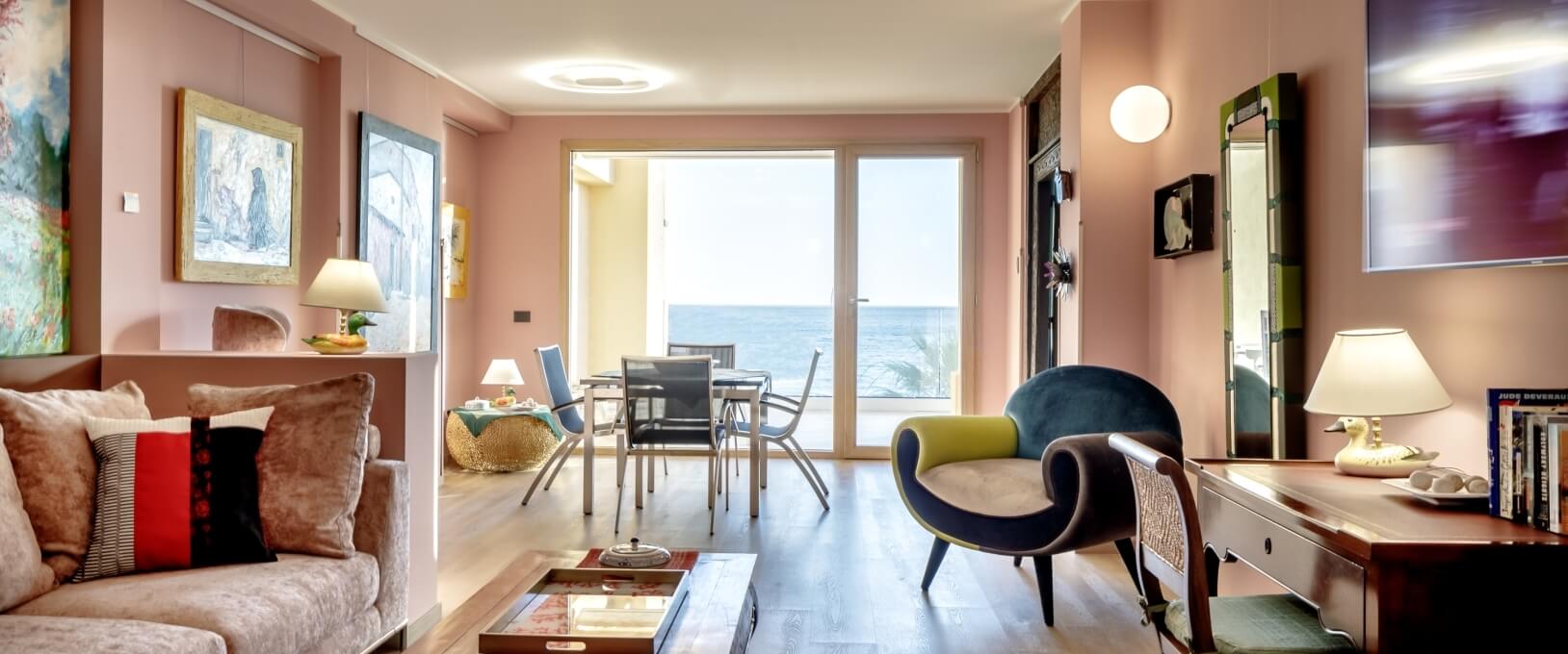 Salon moderne aux murs roses, canapé beige, fauteuil bleu et jaune, vue sur l'océan à travers des portes-fenêtres.