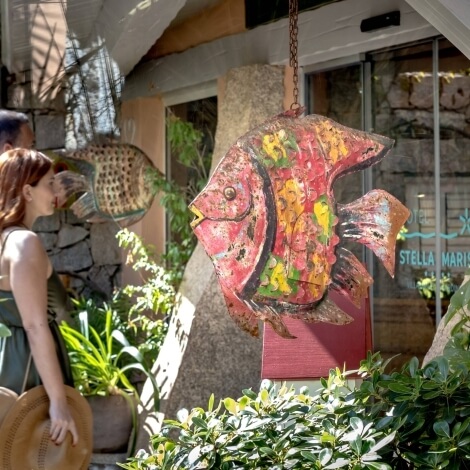 A woman in a green dress and a man in a gray shirt stand by a colorful metal fish sculpture outside "STELLA MARIS."