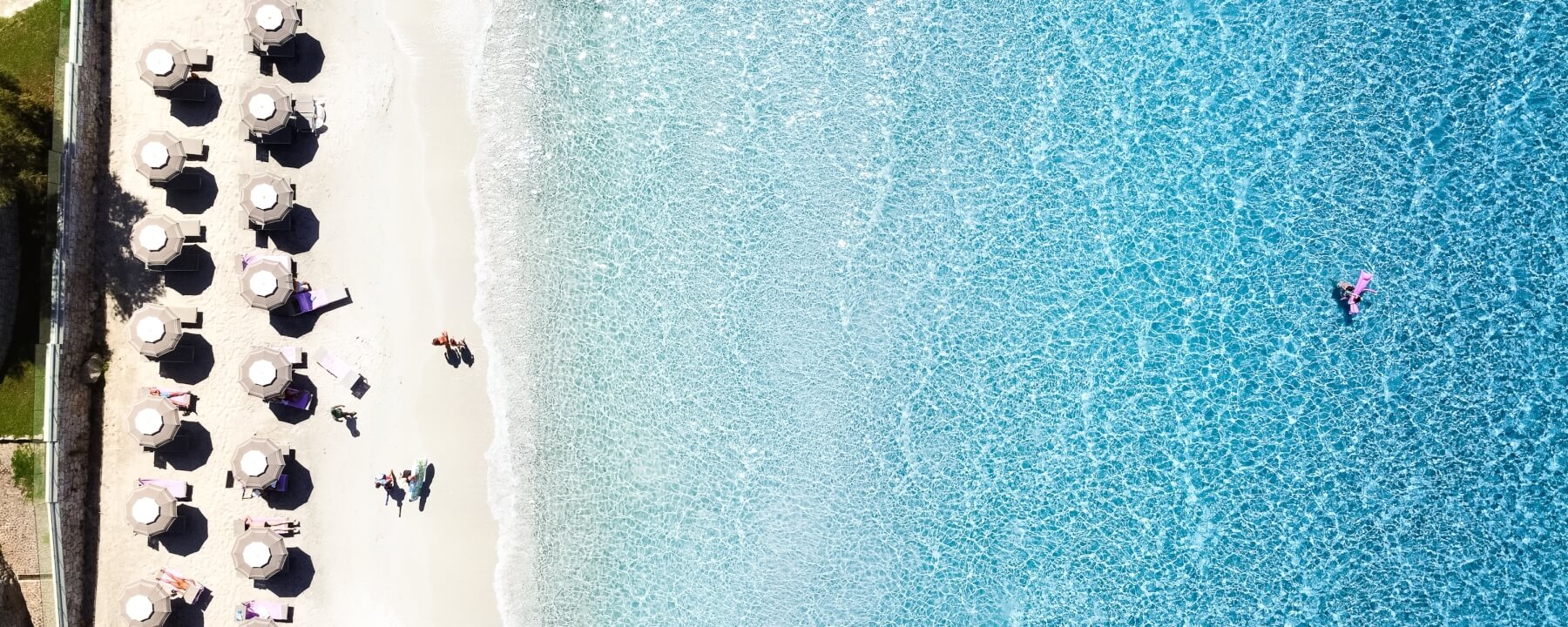 Aerial view of a serene beach with umbrellas, lounge chairs, and people relaxing, alongside clear blue ocean waters.