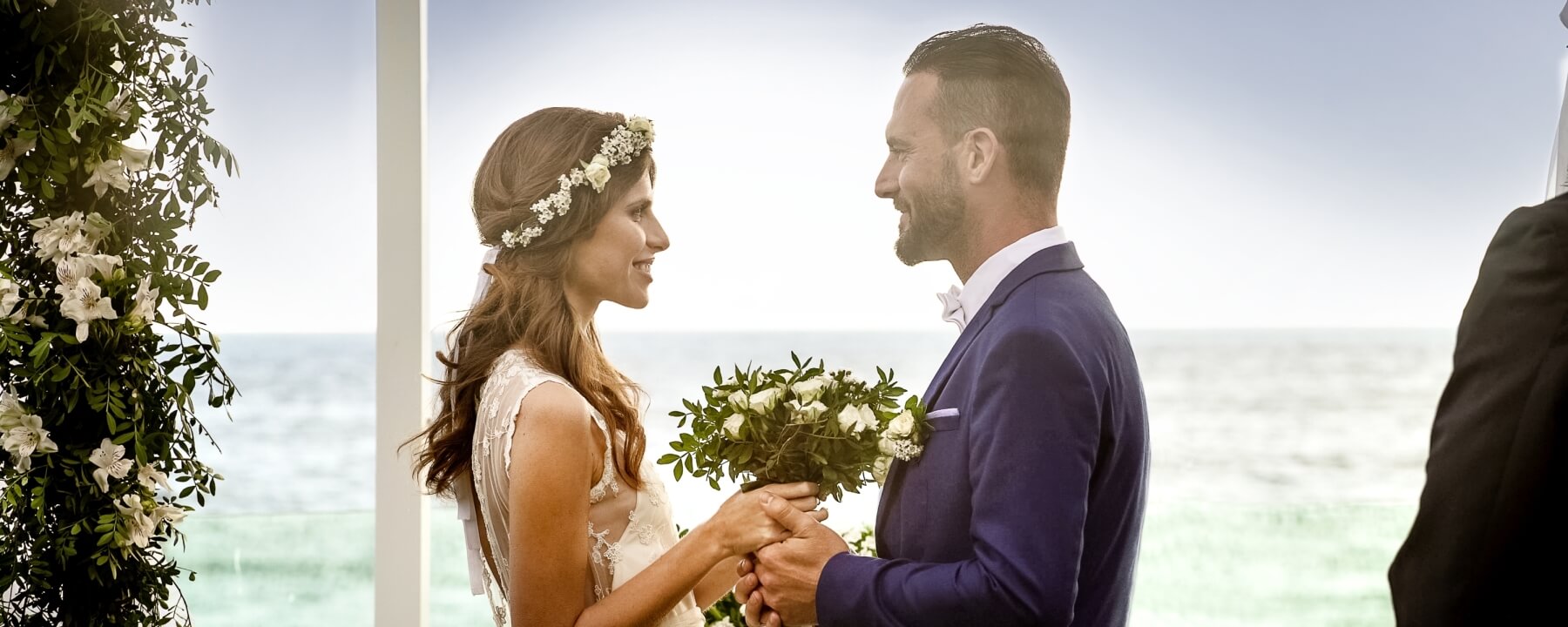 Un couple échange des vœux sur une plage, entouré de verdure, avec l'océan et un ciel bleu en arrière-plan.