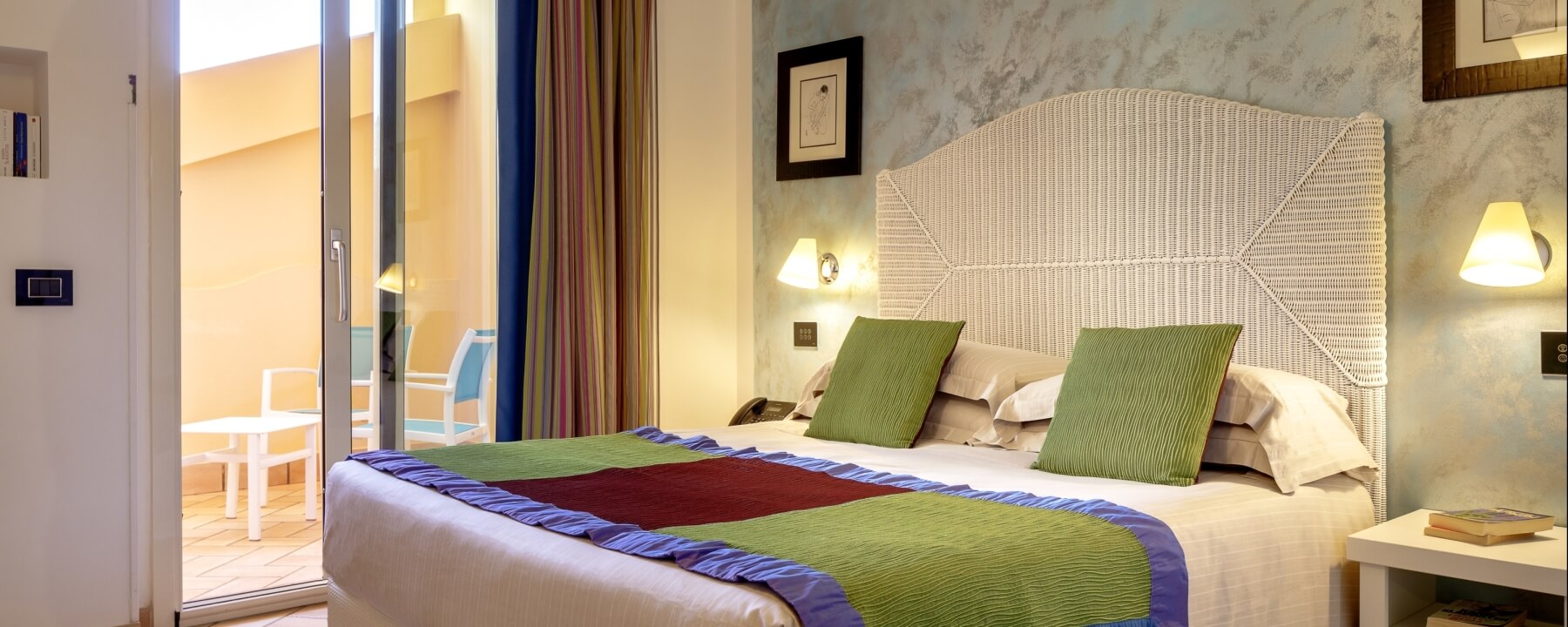 Schlafzimmer mit großem Bett, buntem Quilt, Nachttisch, Lampen und Balkon mit zwei Stühlen, gemütliche Atmosphäre.