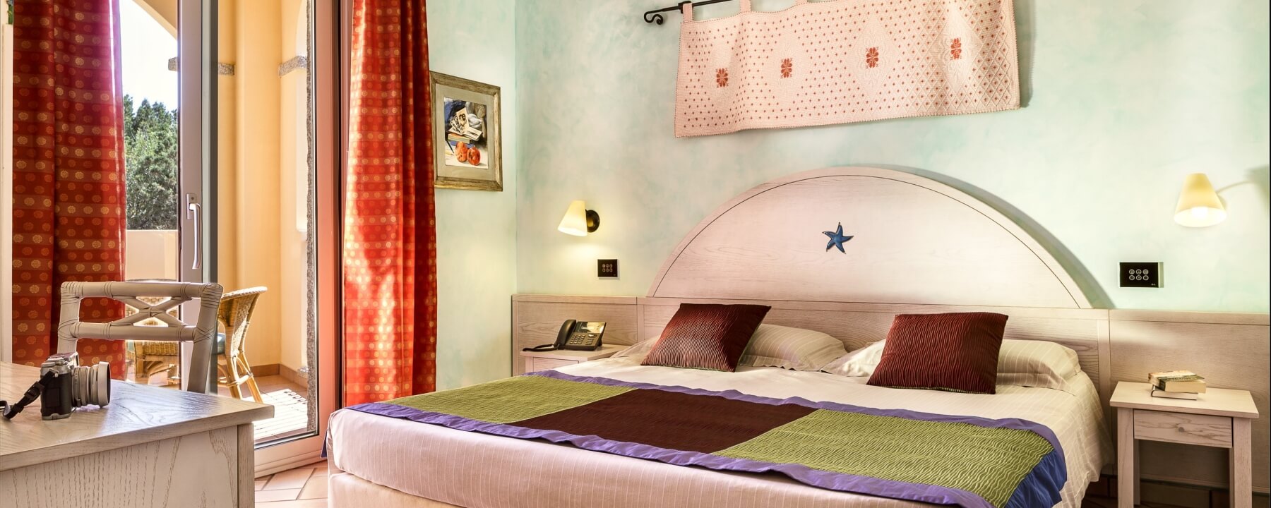 Schlafzimmer mit großem Bett, Nachttischen, Schreibtisch, buntem Quilt und roter Vorhänge vor einem Balkon.