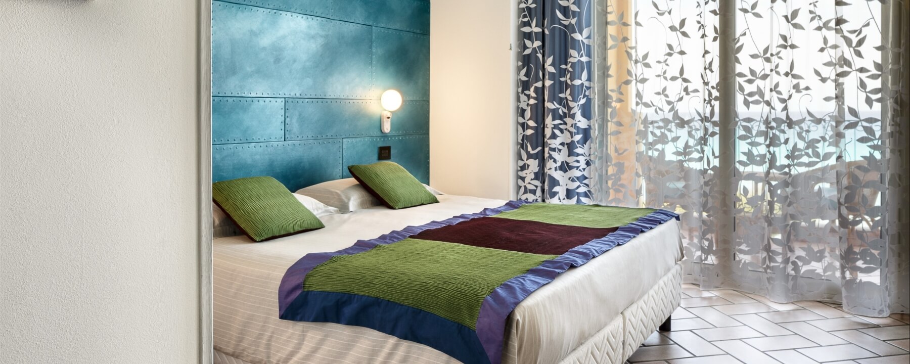 Chambre avec un lit à la tête contre un mur bleu, rideaux transparents et sol en carrelage blanc.