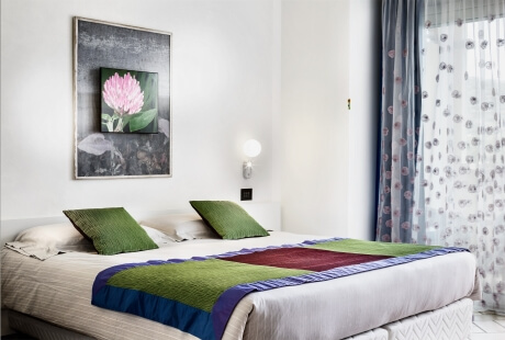 Hotel room with bed, green pillows, floral artwork, and patterned curtains.