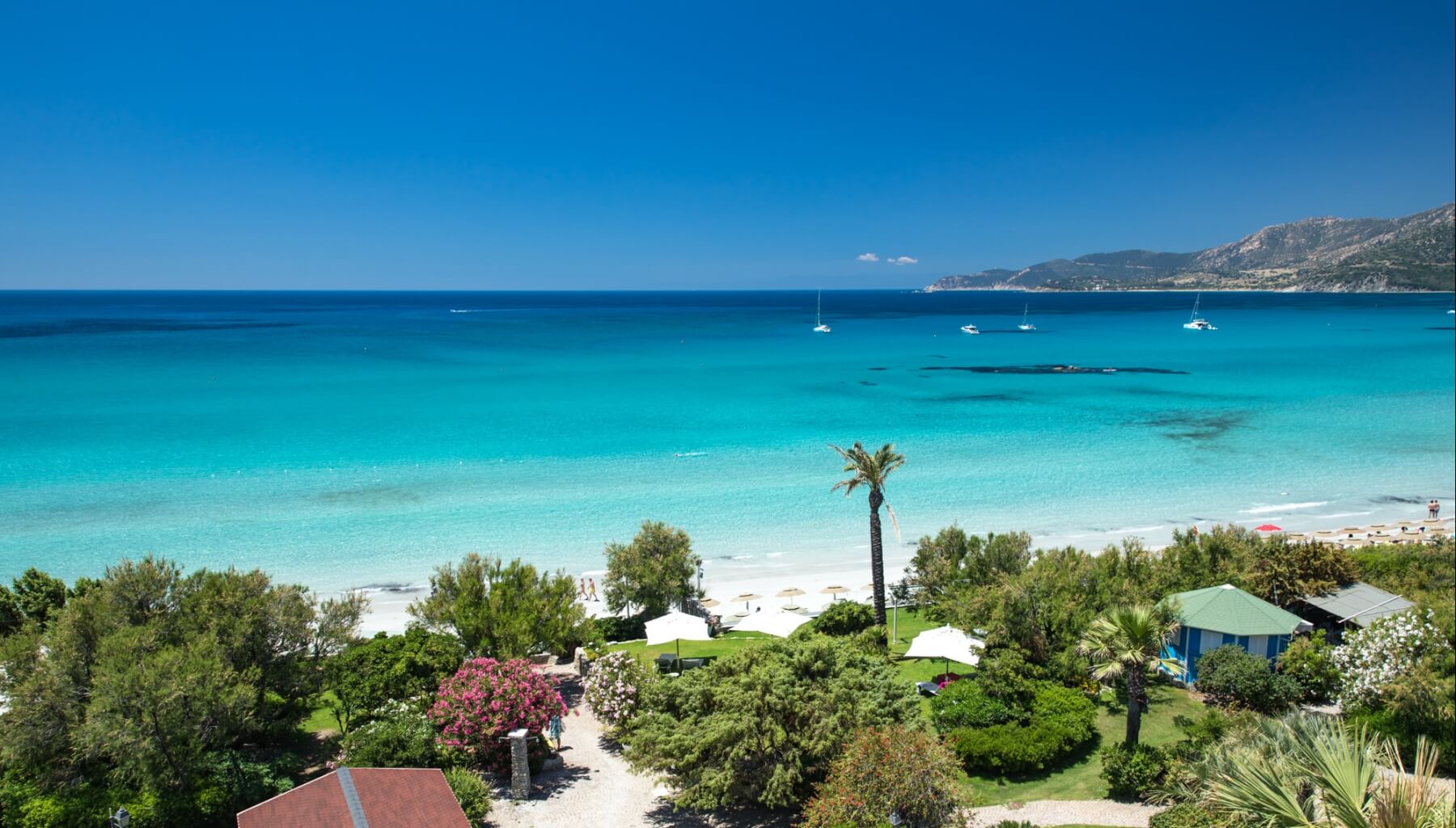 Serene beach scene with turquoise sea, boats, white umbrellas, lush greenery, and distant mountains under a blue sky.