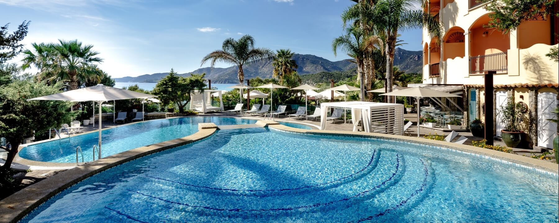 Luxurious outdoor pool scene with a large curved pool, lounge chairs, umbrellas, lush greenery, and mountains under a blue sky.