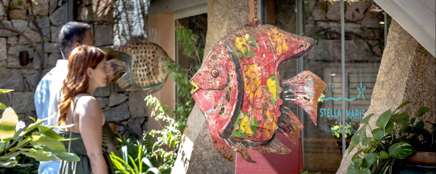 Coppia davanti a un hotel con sculture di pesci, piante in vaso e scritta "HOTEL STELLA MARIS" sullo sfondo.