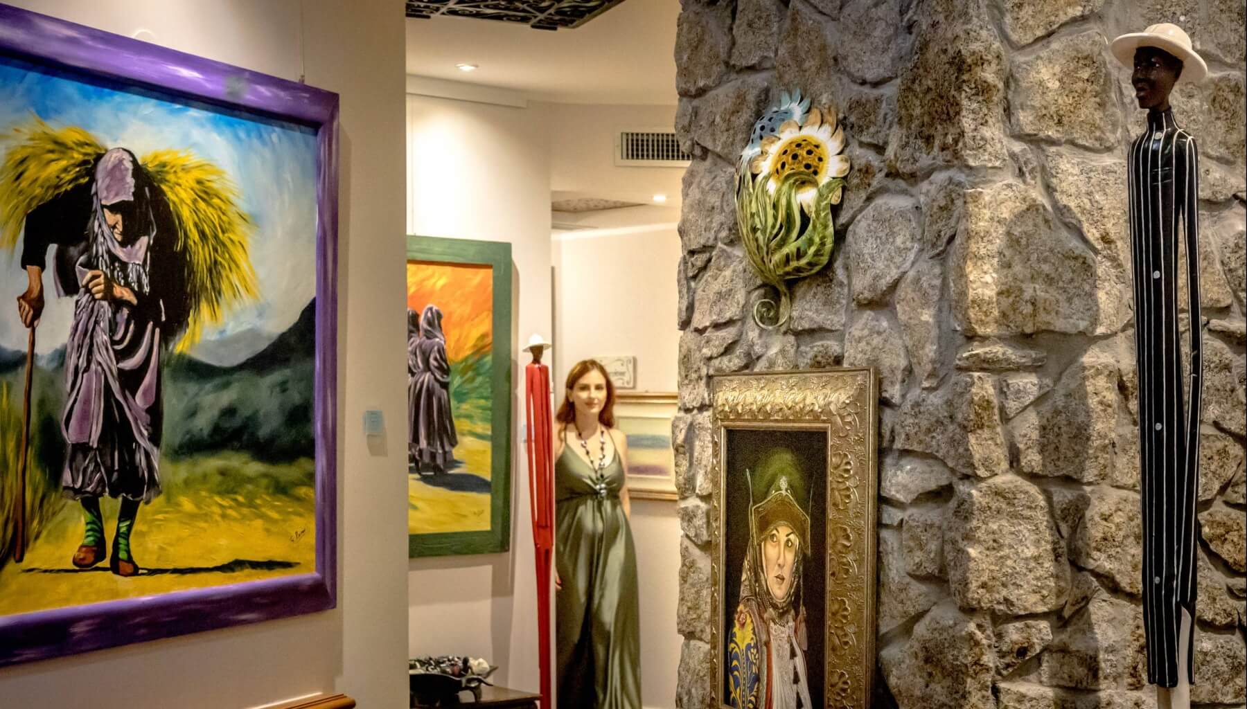 A woman in a green dress stands in an art exhibition with various paintings and sculptures in a high-ceilinged space.