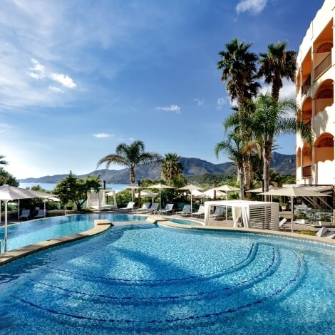 Serene kidney-shaped pool with ripples, surrounded by umbrellas, chairs, palm trees, and a multi-level building under a blue sky.