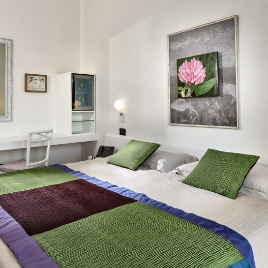 Camera con letto bianco, cuscini verdi e maroon, arte floreale rosa, scrivania e lampada calda.