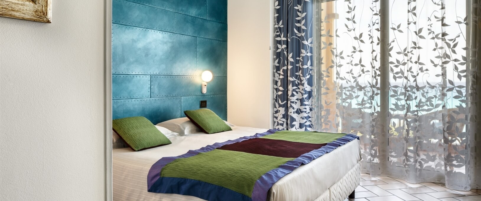 Chambre paisible avec un lit blanc, coussins verts, mur bleu, fenêtre avec rideaux légers et éclairage doux.