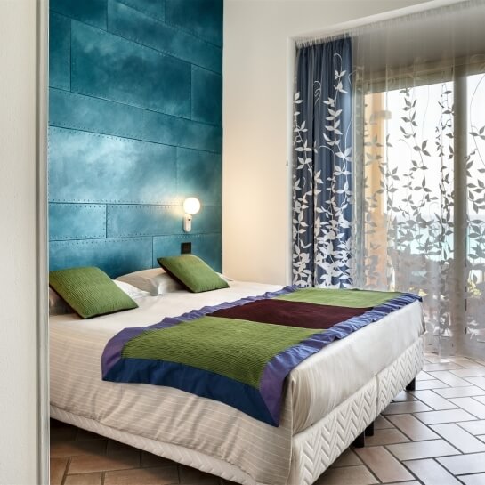 Camera serena con letto matrimoniale, coperta bianca decorata, cuscini verdi e bianchi, parete blu e luce calda.