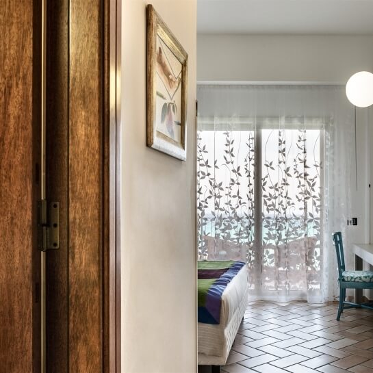 Interno sereno con porta in legno, pareti bianche, letto elegante, sedia teal e luce calda, creando un'atmosfera accogliente.