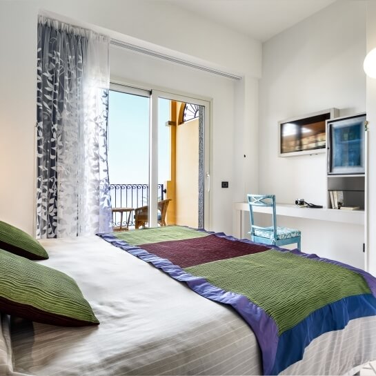 Camera serena con letto, scrivania e TV, grande finestra con vista mare e tende bianche che illuminano l'ambiente.
