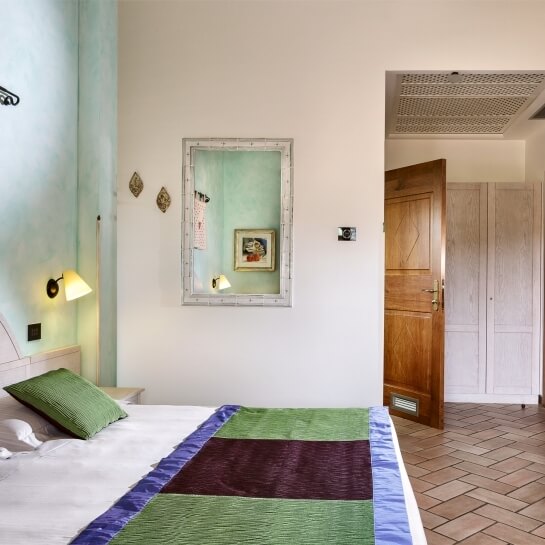 Camera da letto con letto bianco, cuscini verdi, lampada, specchio e porta aperta verso un'altra stanza.