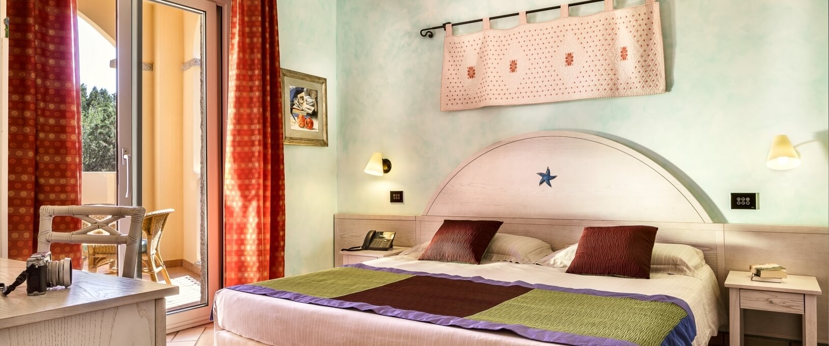 Chambre sereine avec lit, couvre-lit blanc, tapis rose, portes ouvertes sur un patio avec chaises en osier et table.