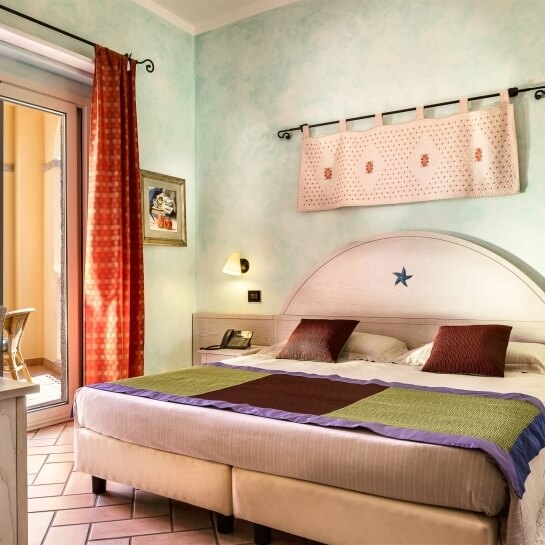 Camera con letto centrale, testiera bianca, coperte colorate, tende beige e rosse, e pareti blu-verdi.