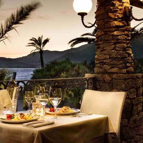 A beautifully set dinner table on a patio, overlooking water and mountains, illuminated by sunset and surrounded by greenery.