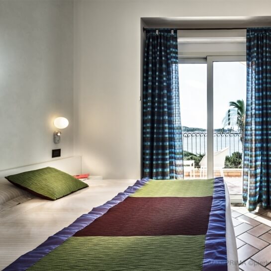 Una stanza serena con un letto, una porta aperta su un balcone e una vista panoramica di alberi e cielo nuvoloso.