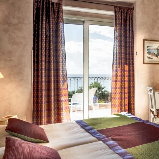 Una stanza serena con vista mare, decorata con tende colorate e un letto bianco, trasmette tranquillità e relax.