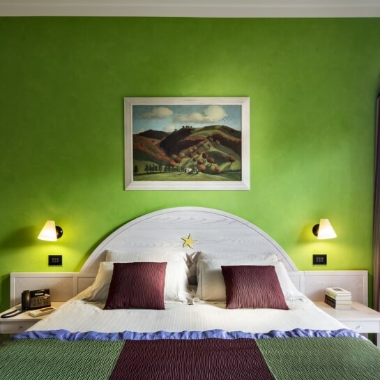 Lit large avec tête de lit blanche, oreillers rouges, tables de nuit blanches, peinture de paysage, mur vert et tapis.