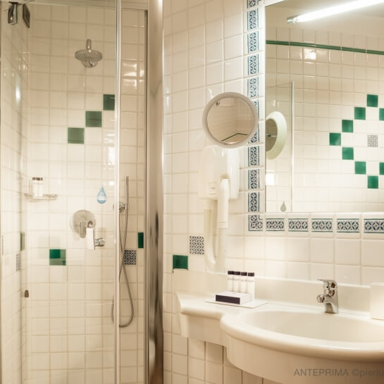 Scena di un bagno con doccia in vetro, lavabo bianco e piastrelle bianche e verdi, con dettagli decorativi.
